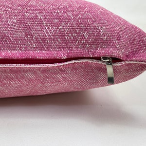 May include: A pink pillow with a white zipper. The pillow is made of a textured fabric.