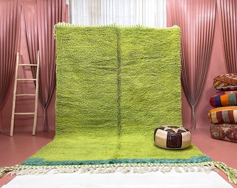 May include: A green shag rug with a fringe border. The rug is laid out on a light pink floor with a wooden ladder and a decorative stool in the background.