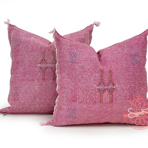 May include: Two pink throw pillows with embroidered geometric patterns. The pillows have tassels on the corners and are made of a soft, textured fabric.