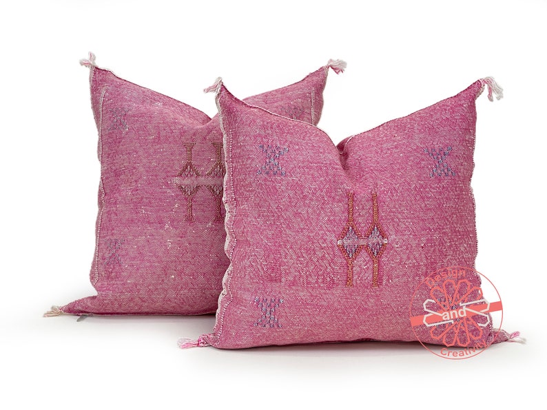 May include: Two pink throw pillows with embroidered geometric patterns. The pillows have tassels on the corners and are made of a soft, textured fabric.