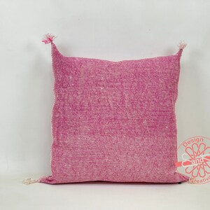 May include: A pink throw pillow with a textured woven fabric and white fringe tassels on the corners.
