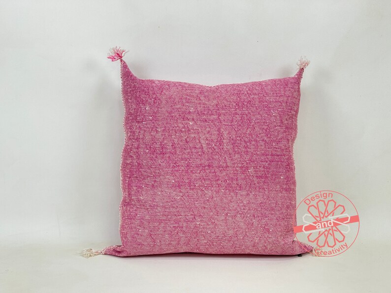 May include: A pink throw pillow with a textured woven fabric and white fringe tassels on the corners.