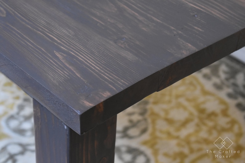 May include: Close-up of a dark stained wooden table top with visible wood grain. The table has a square top and a single leg visible in the image.