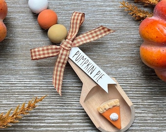 May include: A wooden scoop ornament with a brown and white gingham bow, three beads in white, orange, and tan, and a white tag that says "Pumpkin Pie". The scoop has a small orange and brown pie slice inside.
