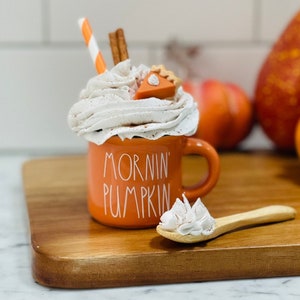 May include: Orange ceramic mug with the text "Mornin' Pumpkin" on it. The mug is filled with whipped cream and a miniature pumpkin pie slice. A wooden spoon with whipped cream sits on a wooden cutting board.