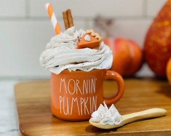 May include: Orange ceramic mug with the text "Mornin' Pumpkin" on it. The mug is filled with whipped cream and a miniature pumpkin pie slice. A wooden spoon with whipped cream sits on a wooden cutting board.