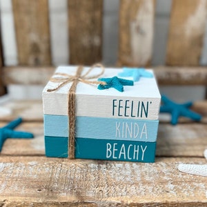 May include: Three stacked wooden blocks painted in shades of blue and white. The top block reads "Feelin' Kinda Beachy" in white lettering.