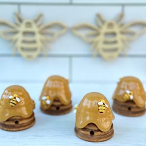 May include: Four small ceramic honey pot candle holders with bee accents. The candle holders are glazed in a honey color and have a textured surface.