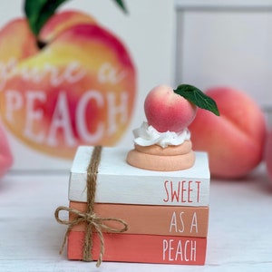 May include: A stack of three small wooden books with peach-colored, orange, and white painted covers. The books are tied together with twine and have the words "Sweet as a Peach" written on them. A peach-colored faux peach with a green leaf sits on top of the books.