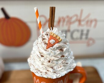 May include: An orange mug with the words "MORNING PUMPKIN" in white, topped with whipped cream, a pie slice, cinnamon, a straw, and a cinnamon stick. A wooden spoon with a whipped cream topping rests on the side. The mug sits on a wooden surface.