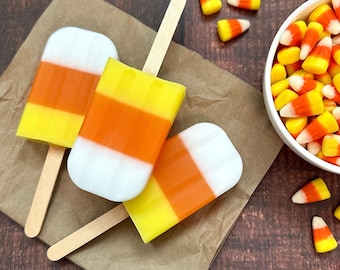 Puede incluir: Tres paletas de hielo naranja, blanco y amarillo con diseños de maíz dulce sobre una bolsa de papel marrón. Las paletas de hielo están en palos de madera. Un tazón blanco lleno de maíz dulce está en el fondo.