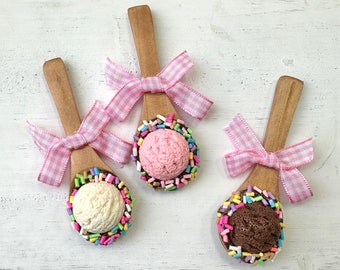 Puede incluir: Tres cucharas de madera con lazos rosas y espolvoreadas con coloridos sprinkles alrededor de los bordes. Cada cuchara contiene un sabor diferente de helado: vainilla, fresa y chocolate.