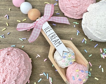 Puede incluir: Un adorno de cuchara de helado de madera con un lazo de vichy rosa y blanco y las palabras "ICE CREAM" en una etiqueta blanca. El adorno está rodeado de bolas de helado rosa y blanco y espolvoreos coloridos.