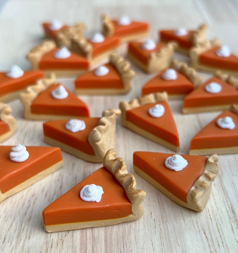 May include: A collection of miniature pumpkin pie slices with a dollop of whipped cream on top. The pies are made of polymer clay and are a light orange color with a brown crust.