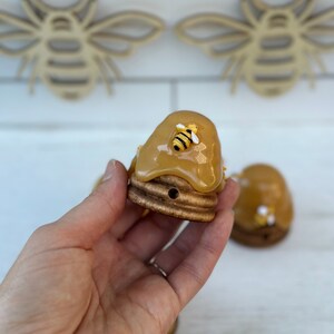 May include: A small wooden beehive with a yellow honey-like glaze and a small yellow bee figurine on top. The beehive is sitting on a hand.