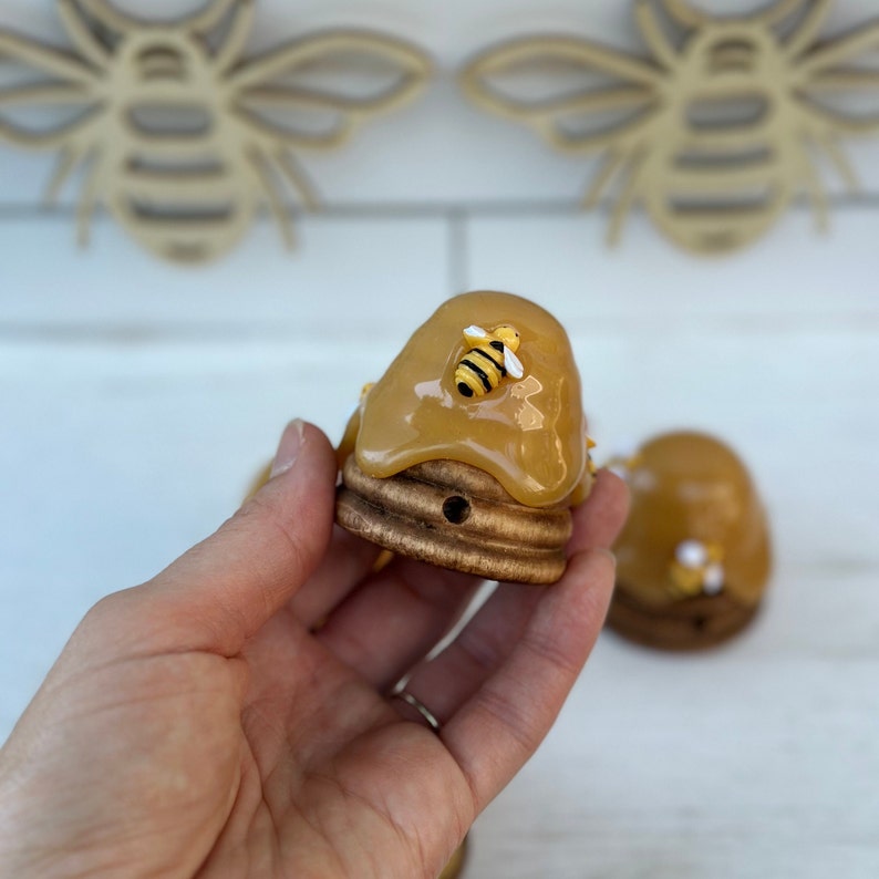 May include: A small wooden beehive with a yellow honey-like glaze and a small yellow bee figurine on top. The beehive is sitting on a hand.