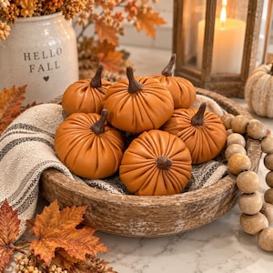 May include: A collection of five orange fabric pumpkins with dark brown stems arranged in a round wooden bowl. The pumpkins feature gathered pleats and a soft texture. A striped fabric cloth lines the bowl, and a string of wooden beads rests nearby.
