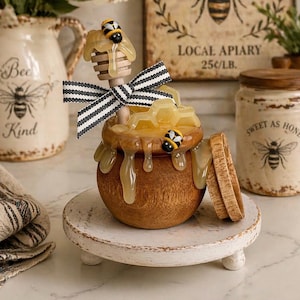 May include: A wooden honey pot with a lid, honey dipper, and decorative bees. The pot sits on a white, round, pedestal stand. A black and white striped ribbon is tied around the honey dipper. The jar is filled with honey.