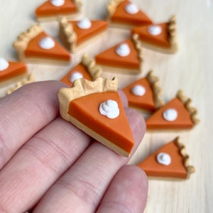 May include: A collection of miniature pumpkin pie slices made of polymer clay. Each slice is decorated with a dollop of white frosting. The slices are arranged on a wooden surface.