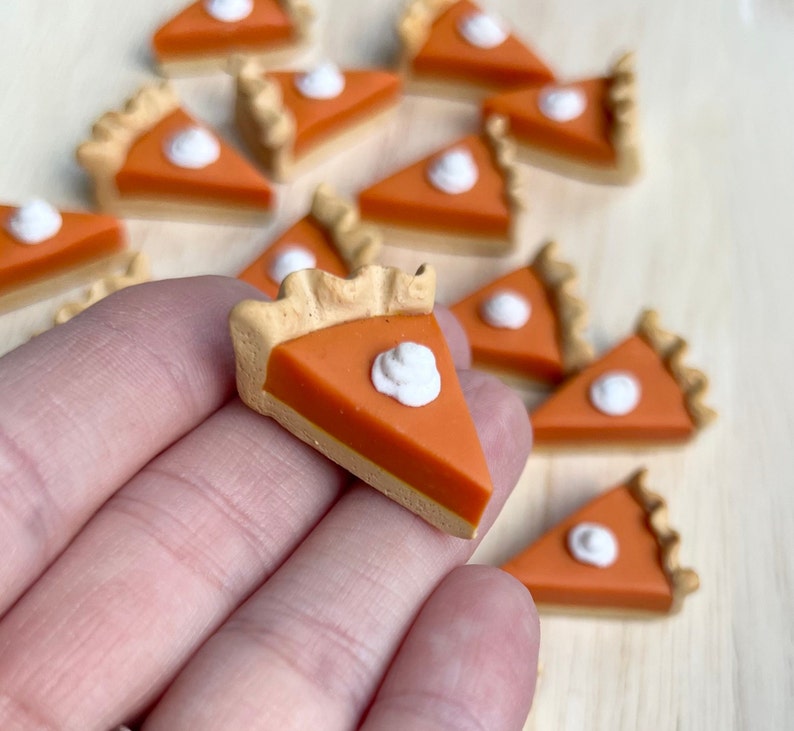 May include: A collection of miniature pumpkin pie slices made of polymer clay. Each slice is decorated with a dollop of white frosting. The slices are arranged on a wooden surface.