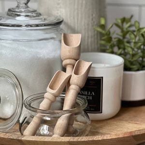 May include: Two wooden scoops with a rounded handle and a scoop at the end. The scoops are sitting in a clear glass jar on a wooden tray. There is a white candle and a glass jar with a lid in the background.