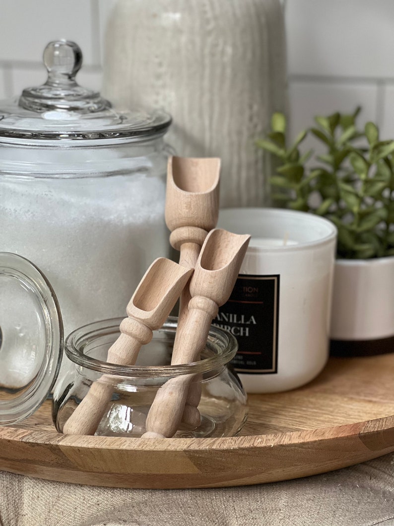 May include: Two wooden scoops with a rounded handle and a scoop at the end. The scoops are sitting in a clear glass jar on a wooden tray. There is a white candle and a glass jar with a lid in the background.