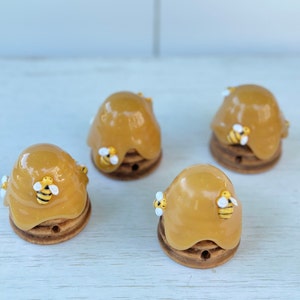 May include: Four small ceramic beehive-shaped ornaments with yellow and brown glaze and painted bee details. The beehives are arranged on a white surface.