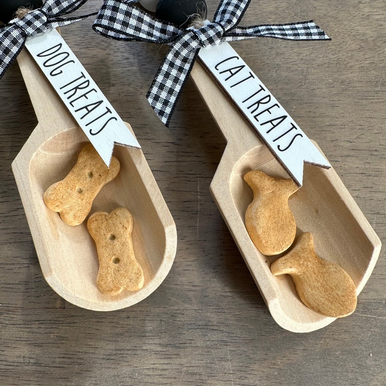 May include: Two wooden scoops with black and white checkered ribbon bows. The scoops are labeled "Dog Treats" and "Cat Treats". The scoop labeled "Dog Treats" contains two bone-shaped dog treats. The scoop labeled "Cat Treats" contains two fish-shaped cat treats.