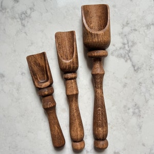 May include: Three wooden scoops of varying sizes, all with a dark brown stain. The scoops have a rounded, spoon-like shape at the top and a handle that tapers to a point.