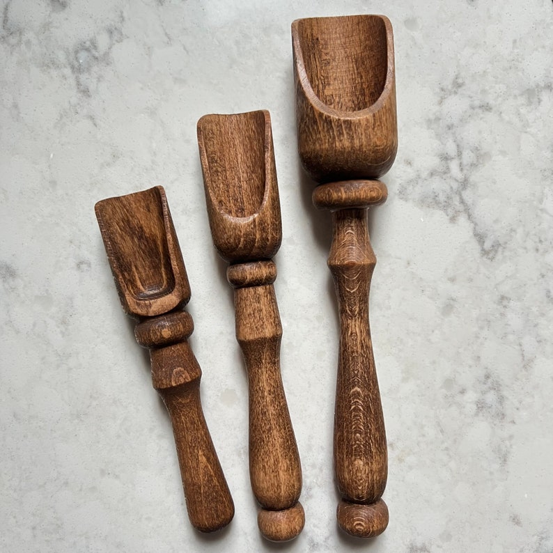 May include: Three wooden scoops of varying sizes, all with a dark brown stain. The scoops have a rounded, spoon-like shape at the top and a handle that tapers to a point.