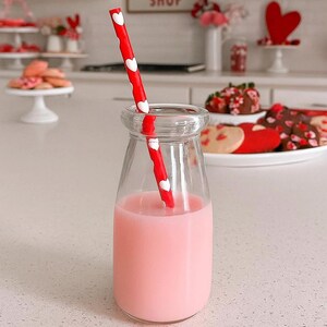 May include: A clear glass milk bottle filled with pink liquid and a red and white heart-patterned straw. The bottle sits on a white surface, with heart-shaped cookies and chocolate-covered strawberries in the background. The image has a Valentine's Day theme.