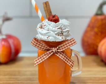 May include: A glass jar with a handle filled with a pumpkin spice latte. The latte is topped with whipped cream, a cinnamon stick, a red and white striped straw, and a small red apple. The jar is tied with a red and white gingham ribbon.