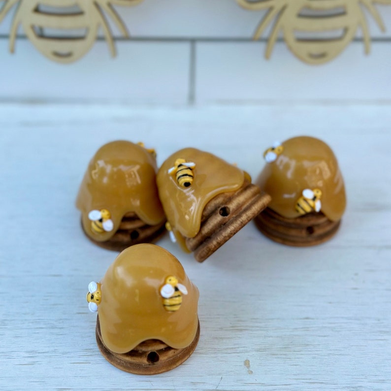 May include: Four small honeycombs with bees on top. The honeycombs are made of wood and have a brown finish. The bees are made of plastic and have yellow and black stripes. The honeycombs are arranged in a semi-circle on a white surface.