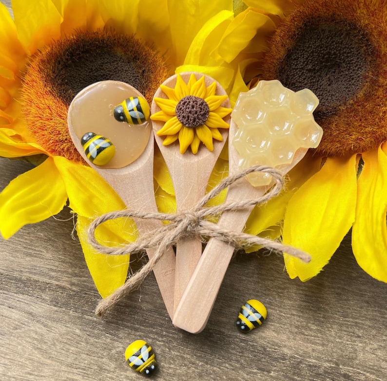 May include: Three wooden spoons with honey and honeycomb on top. The spoons are tied together with twine and decorated with small bee charms. The spoons are surrounded by yellow sunflowers.