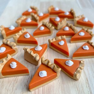 May include: A collection of miniature pumpkin pie slices with a dollop of whipped cream on top. The pies are made of orange and brown fondant and are arranged on a light brown wooden surface.