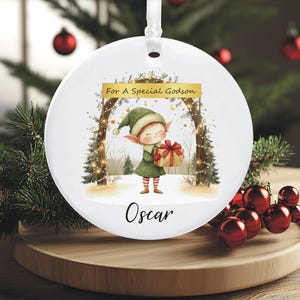 May include: A white Christmas ornament with a ribbon for hanging. The ornament features a watercolor illustration of an elf holding a gift. Text on the ornament reads "For A Special Godson" and the name "Oscar".