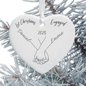 May include: White heart-shaped Christmas ornament with the text "1st Christmas", "Engaged", "Daniel", "Emma", and "2025". The ornament features a drawing of two hands holding each other. A white ribbon is attached for hanging.