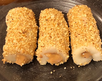 Può includere: Tre rotoli di mochi al burro di arachidi, ciascuno ricoperto da uno strato di arachidi tritate, sono disposti su un piatto di vetro trasparente. I rotoli di mochi sono bianchi e la farcitura al burro di arachidi è visibile attraverso il mochi.