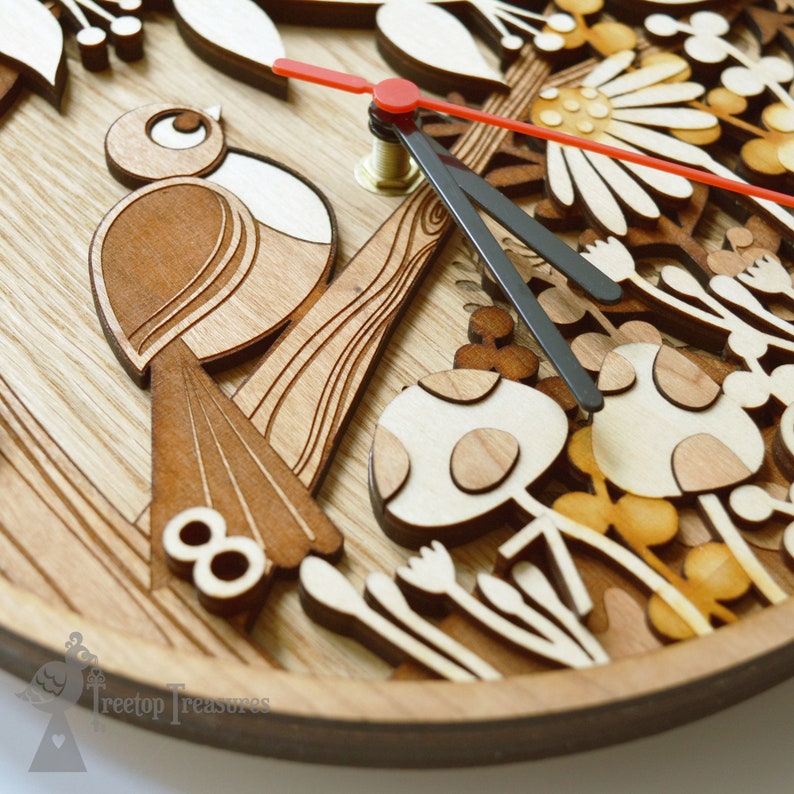 May include: A wooden clock with a bird, mushrooms, and flowers carved into the face. The clock hands are black and red. The clock face is surrounded by a brown wooden frame. The text "Treetop Treasures" is visible at the bottom of the clock.