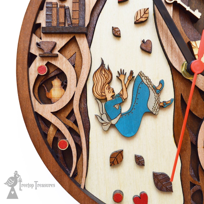 K&ouml;nnte beinhalten: Wanduhr aus Holz mit mehrschichtigem Design zum Thema Alice im Wunderland. Sie zeigt Alice beim Fallen, umgeben von Bl&auml;ttern, mit einem Teeservice und B&uuml;chern. Die Zeiger der Uhr sind rot.