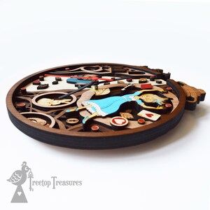 K&ouml;nnte beinhalten: Eine mehrschichtige Holzuhr mit dem Thema Alice im Wunderland. Die Uhr hat einen dunkelbraunen Rahmen und einen hellbraunen und wei&szlig;en Innenraum. Das Design umfasst Alice in einem blauen Kleid, Spielkarten und ein wei&szlig;es Kaninchen. Die Zeiger sind schwarz und rot.