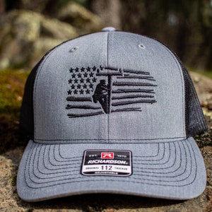 May include: A gray and black trucker hat with a black embroidered American flag featuring a silhouette of a lineman climbing a utility pole. The hat has a black mesh back and a black adjustable strap with a black label that reads "Richardson 112 Adjustable".