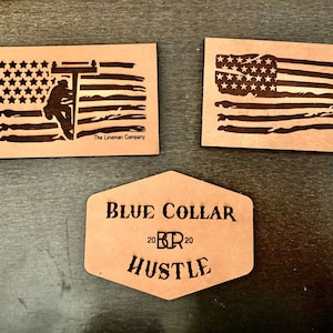 Op de afbeelding: Drie leren patches met een verweerd Amerikaans vlagontwerp. De linker bovenste patch toont een silhouet van een elektricien die aan een hoogspanningslijn werkt. De rechter bovenste patch toont een verweerde Amerikaanse vlag. De onderste patch is zeshoekig en heeft de tekst "BLUE COLLAR 2020 HUSTLE".