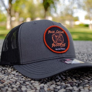 Puede incluir: Una gorra de camionero negra y gris con un parche negro y naranja que dice "Blue Collar Bloodline".