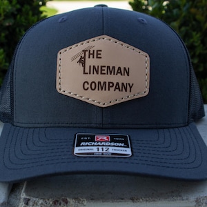 Può includere: Un cappello da camionista grigio scuro con una toppa in pelle marrone che dice "The Lineman Company". La toppa presenta una silhouette di un elettricista che lavora su una linea elettrica. Il cappello è realizzato da Richardson e ha una rete nera sul retro.