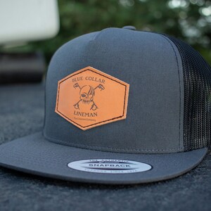 Può includere: Un cappello da camionista grigio e nero con una toppa in pelle marrone che dice "Blue Collar Lineman The Lineman's Company".