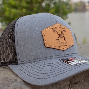 Può includere: Cappellino da camionista grigio e nero con una toppa in pelle marrone che dice "Blue Collar Lineman The Lineman Company".
