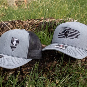 Puede incluir: Dos gorras de camionero grises y negras con un diseño de bandera estadounidense bordado y una silueta de una persona trabajando en una línea eléctrica.