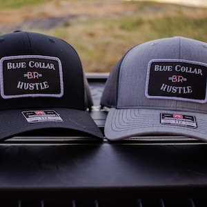 Könnte beinhalten: Zwei Trucker-Caps, eine schwarze und eine graue, mit schwarzen Mesh-Rücken. Jede Cap hat ein rechteckiges Patch mit dem Text "BLUE COLLAR BR20 HUSTLE" in Weiß und Rot. Die Caps sind mit "RICHARDSON 112" versehen.