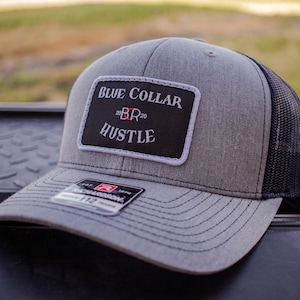 Könnte beinhalten: Eine graue und schwarze Trucker-Kappe mit einem schwarzen Patch, auf dem in weißen Buchstaben "BLUE COLLAR 20 BR 20 HUSTLE" steht.